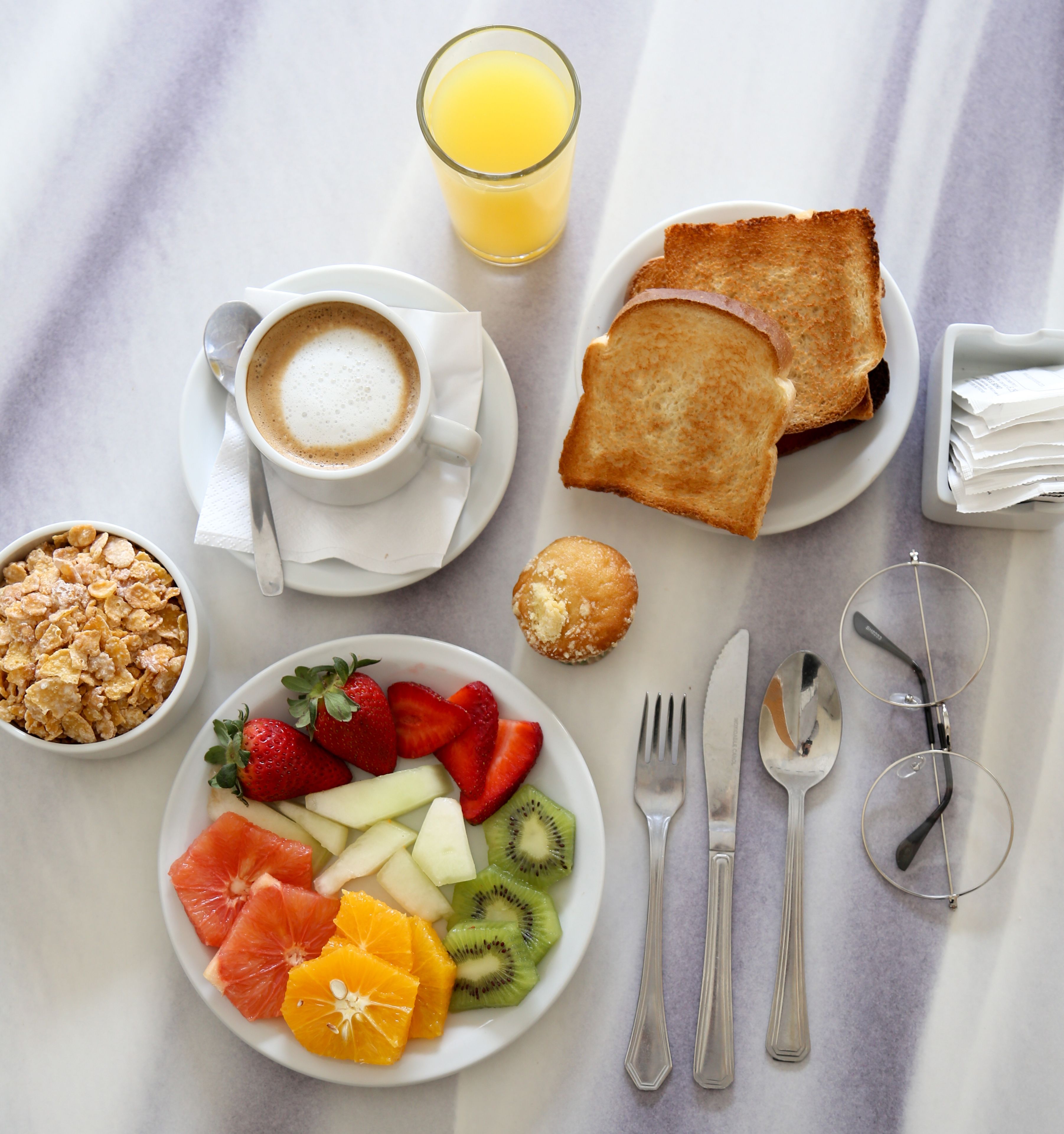 Una mesa con desayuno: tostadas, cereal, fruta, café y jugo.