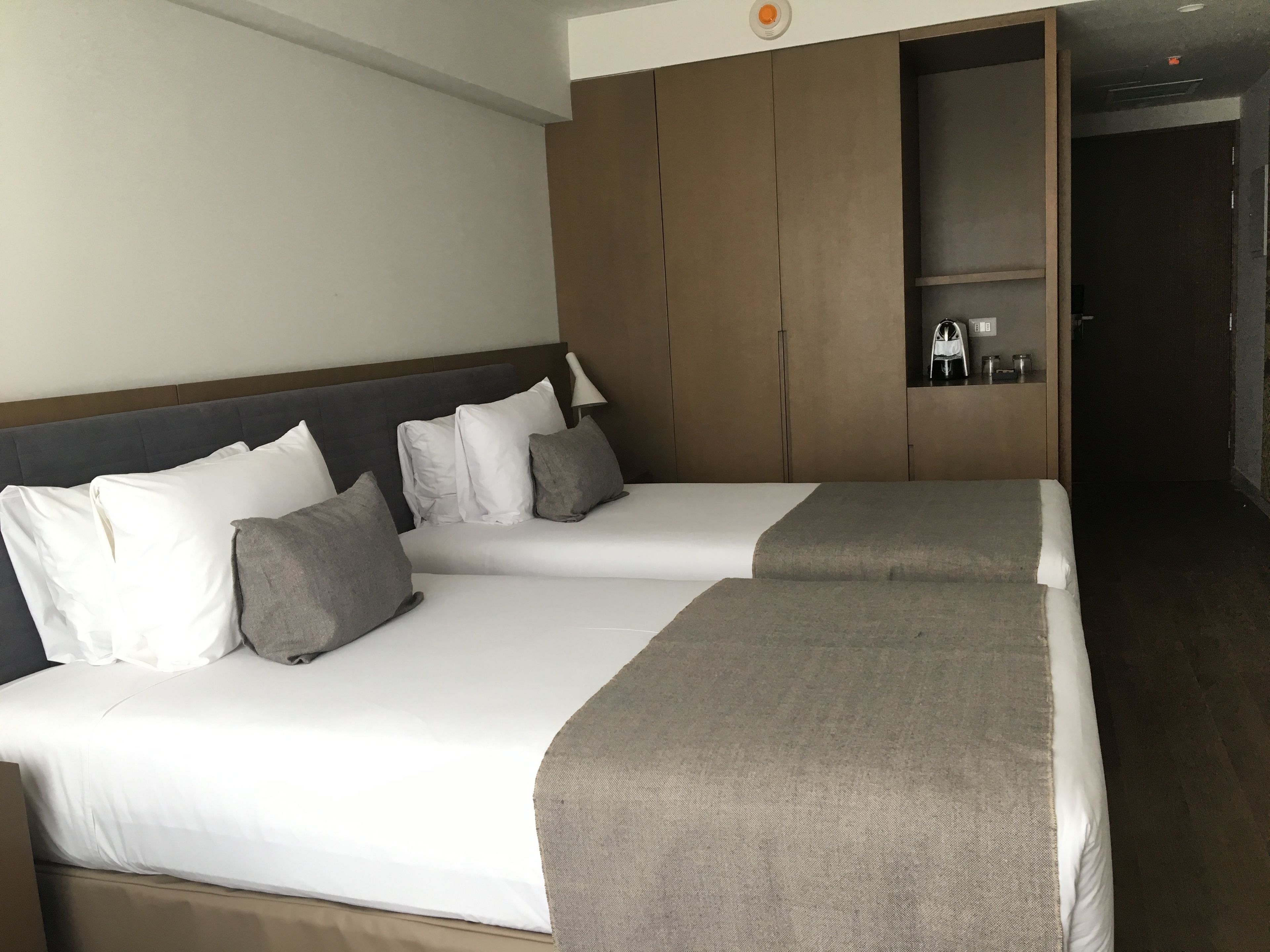A hotel room with two beds, a wooden headboard, a small shelf with toiletries, and a dark wooden door.