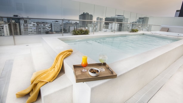 Un espace piscine sur le toit, doté d’une rambarde en verre, offrant une vue sur la ville, avec un plateau en bois sur lequel sont disposés des boissons et des en-cas, et une serviette jaune.