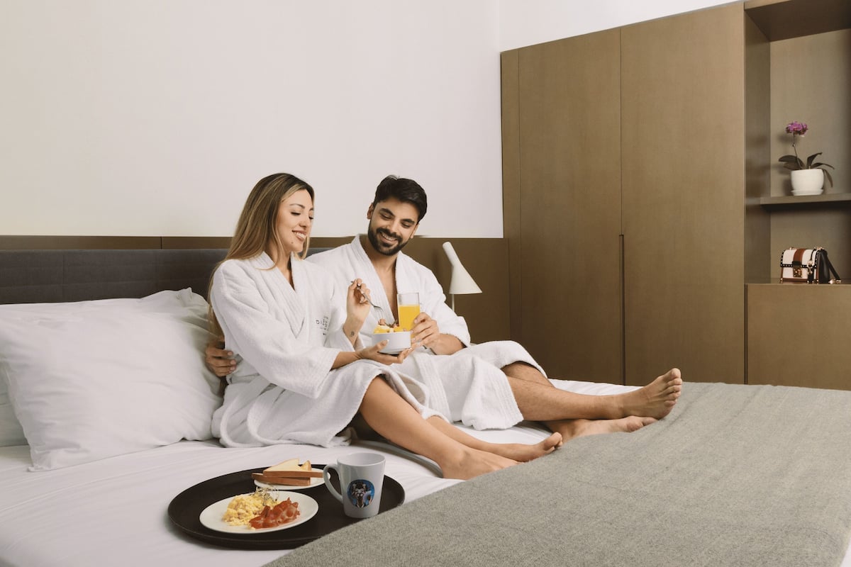 Ein Paar in wei ßen Bademänteln sitzt auf einem Bett mit Frühstücksleckereien, darunter Toast, Eier und Orangensaft.