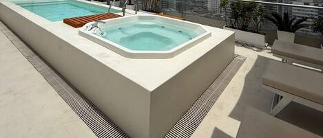 A rooftop pool area with a hot tub and lounge chairs, overlooking a cityscape.