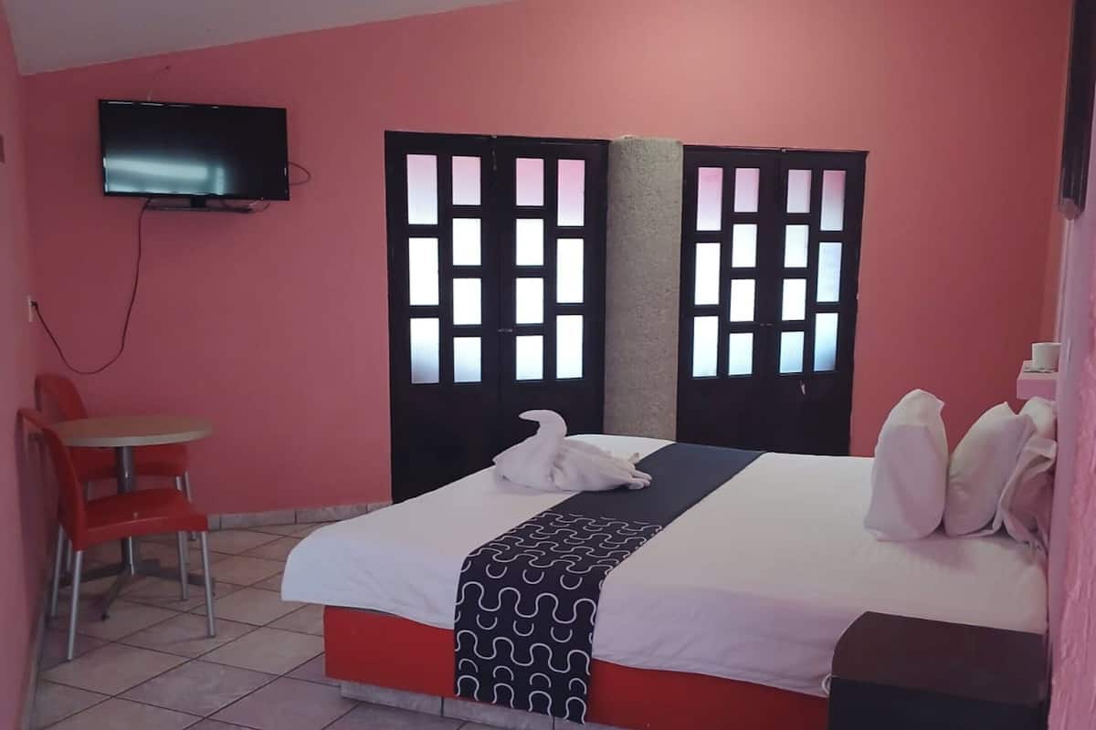 Una habitación con una cama, un televisor fijado a la pared, una silla y una mesita.