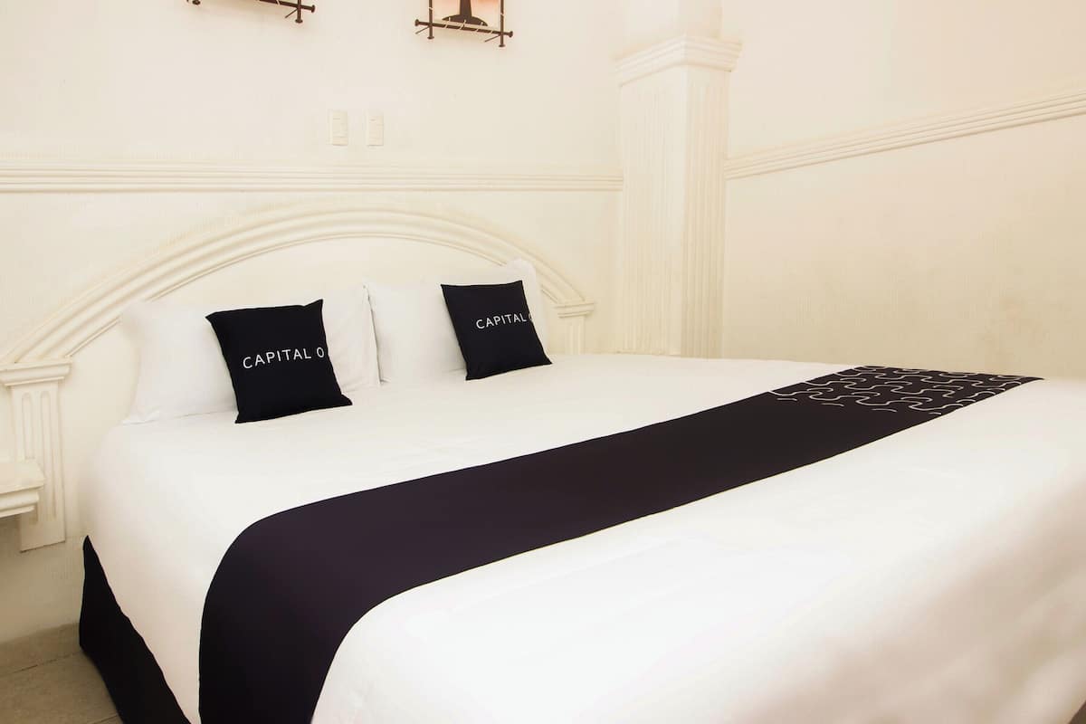 Una cama bien tendida con una colcha blanco y negro, dos almohadillas negras con el texto 'CAPITAL' y un cabecero blanco.