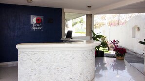 Un área de recepción con suelo de baldosas, un mostrador de recepción, una computadora portátil sobre el mostrador y plantas en macetas.