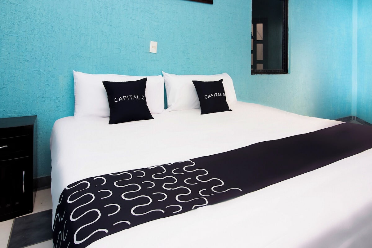Una cama con ropa de cama blanca, almohadas negras y una manta de estilo bicolor, sobre una pared color turquesa.