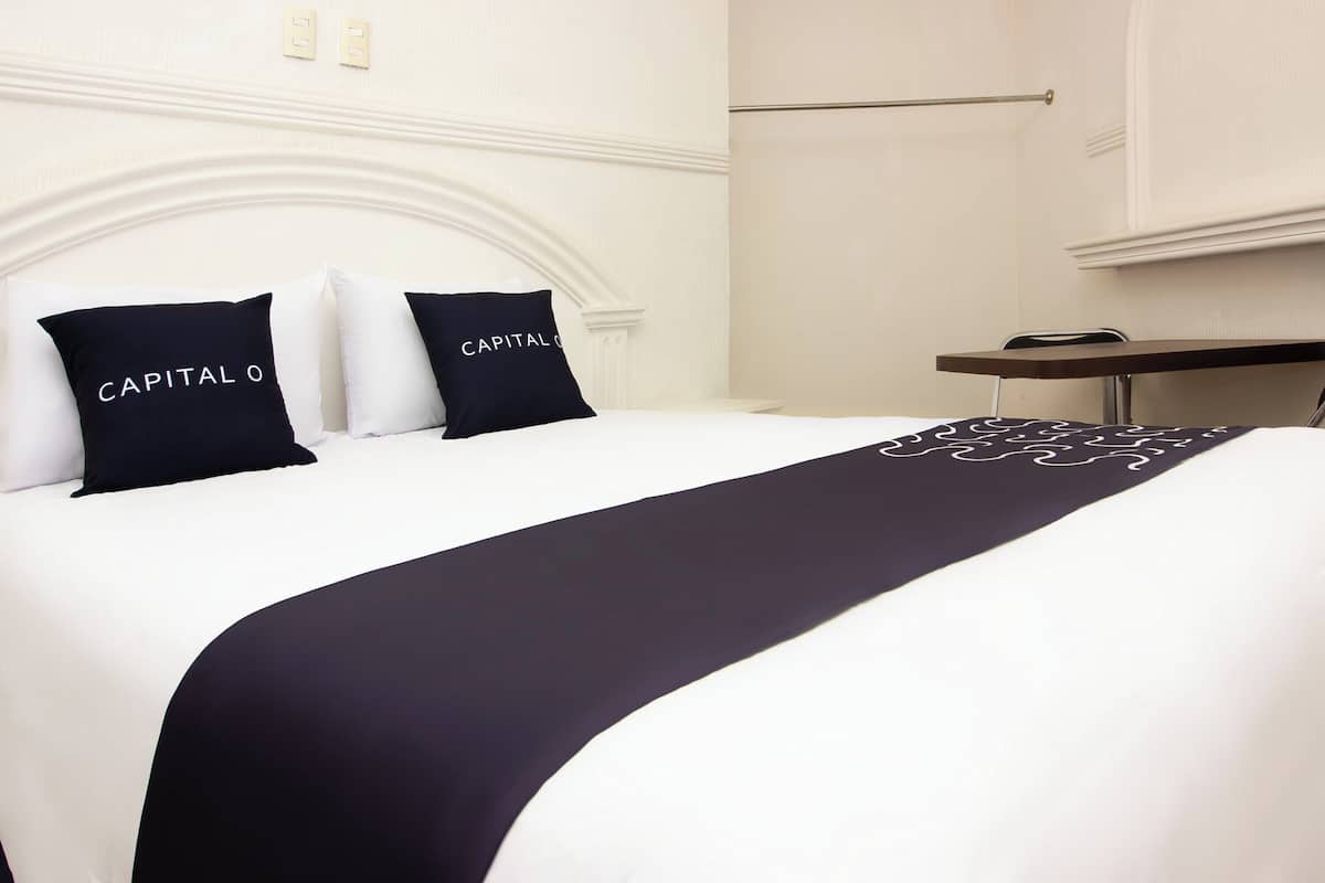 Habitación de hotel con una cama, dos almohadas con el texto 'CAPITAL O', una colcha oscura, una mesita pequeña y una silla.