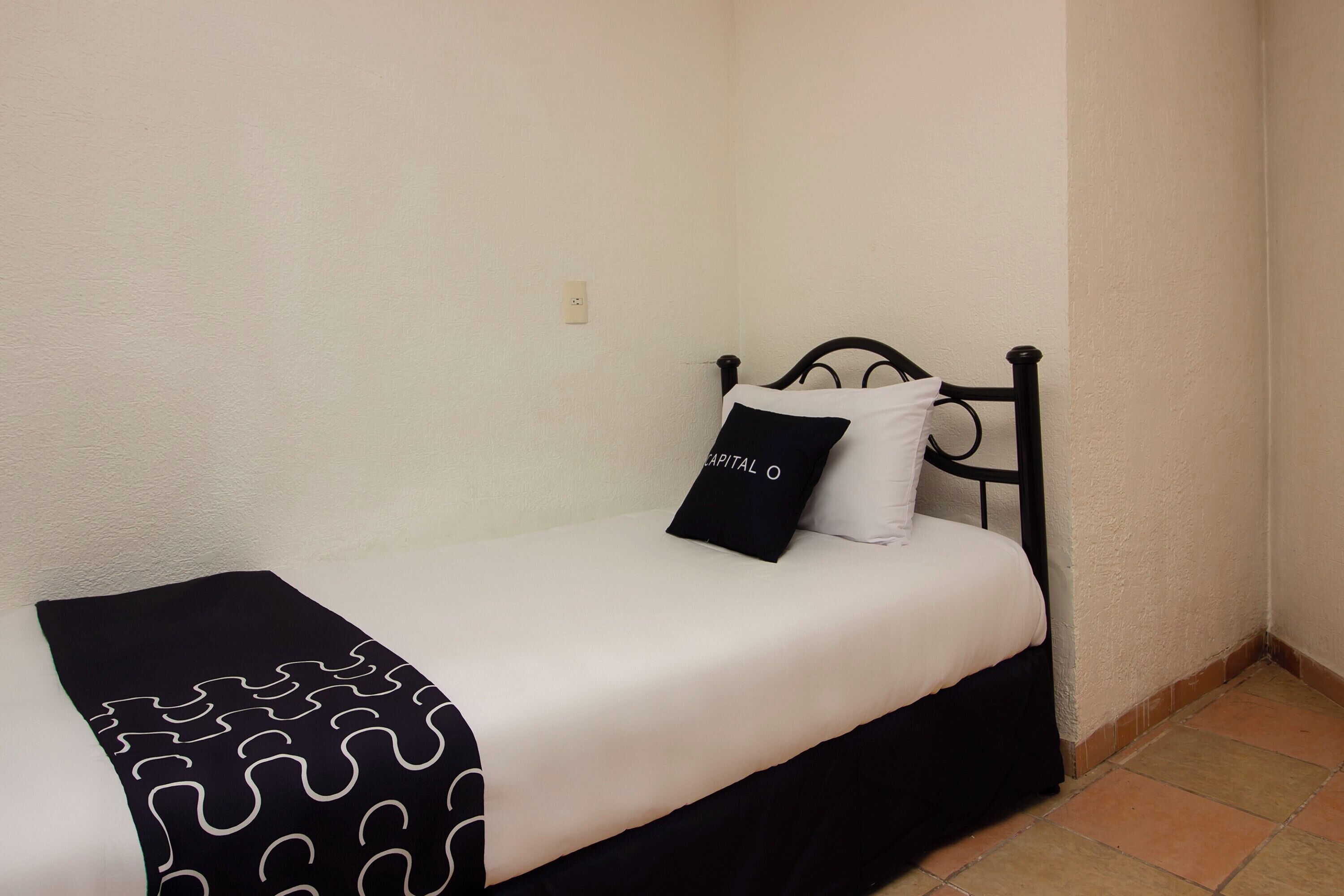 Una cama individual con estructura de metal negro, ropa de cama blanca y almohada negra con texto blanco.