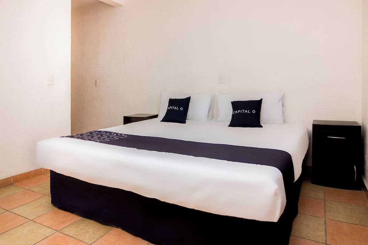 Una cama bien hecha con una colcha blanca y azul oscuro, dos almohadas con el texto 'CAPITAL O' y una mesita de noche negra con cajón.