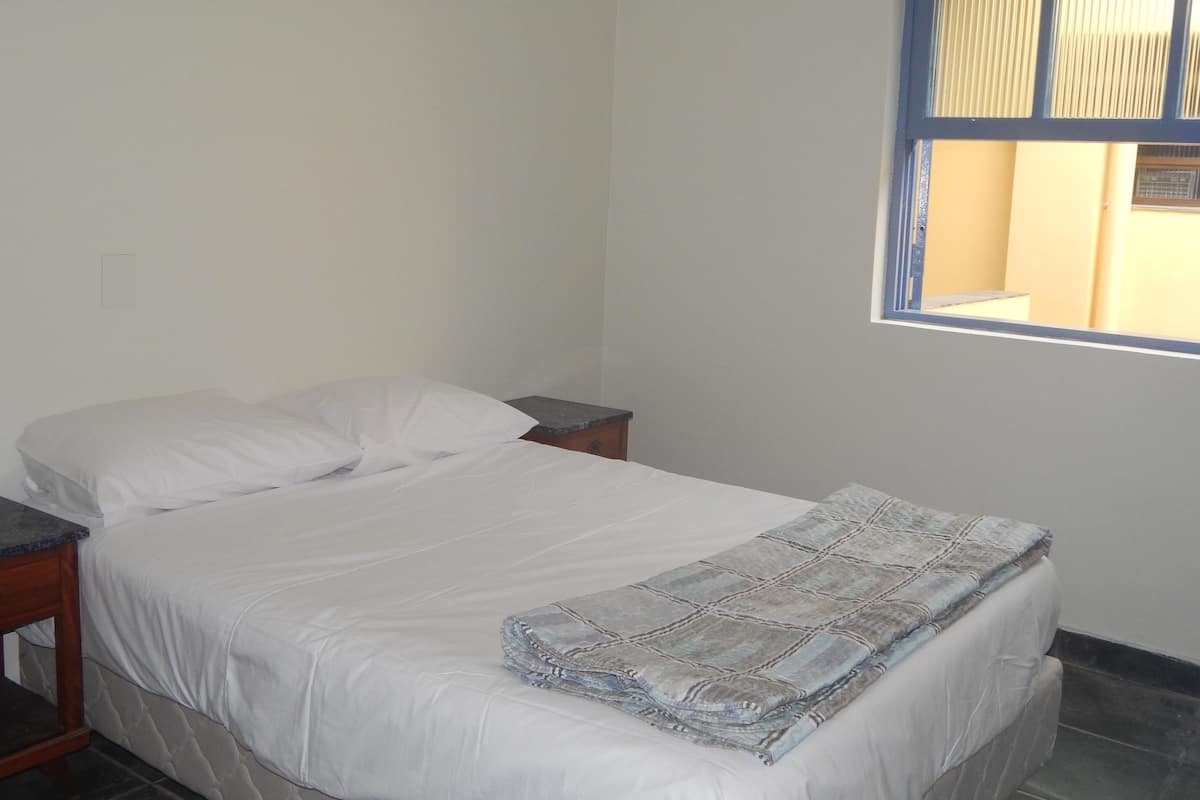 A single bed with white bedding, a small bedside table, and a window with blue shutters.