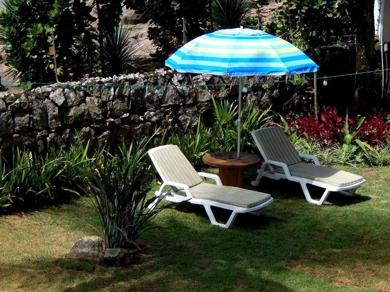 Zwei weiße Lounge-Stühle mit gestreifter Polsterung, ein blau-weiß gestreifter Sonnenschirm und ein kleiner runder Tisch mit Holzplatte.