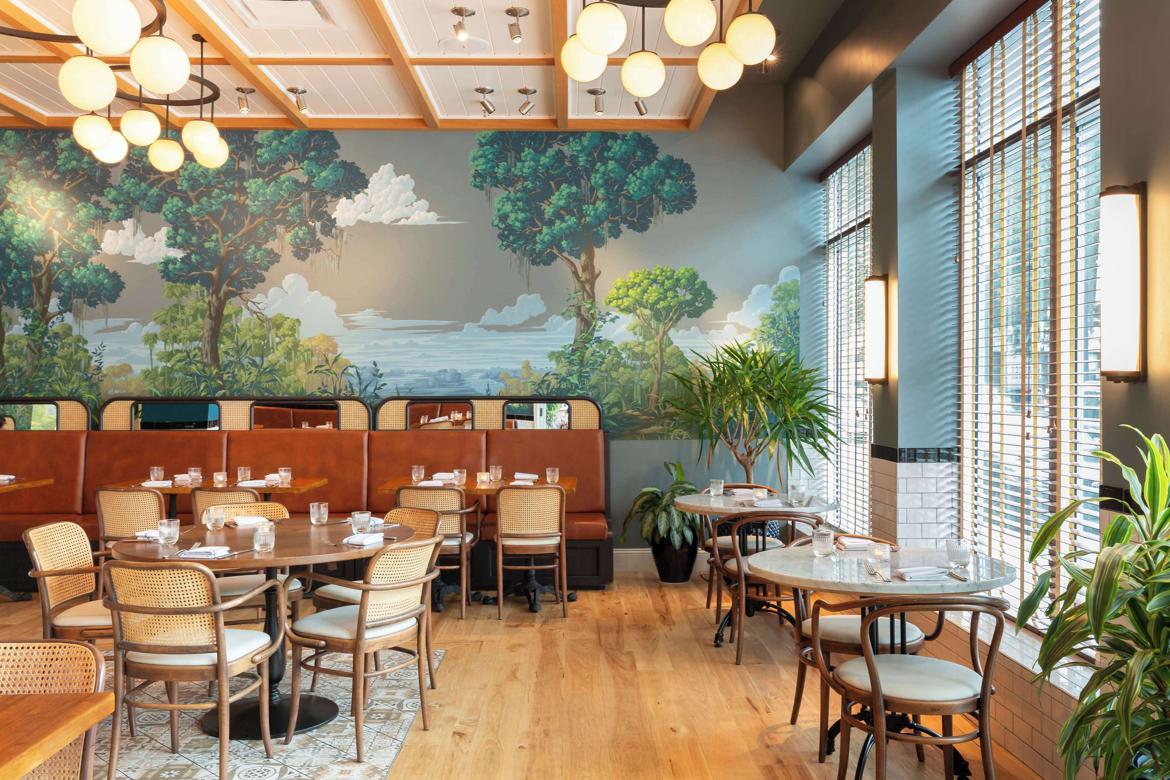 En restaurant med træborde og stole, et muralmaleri af en skov og flod, samt store vinduer med persienner.