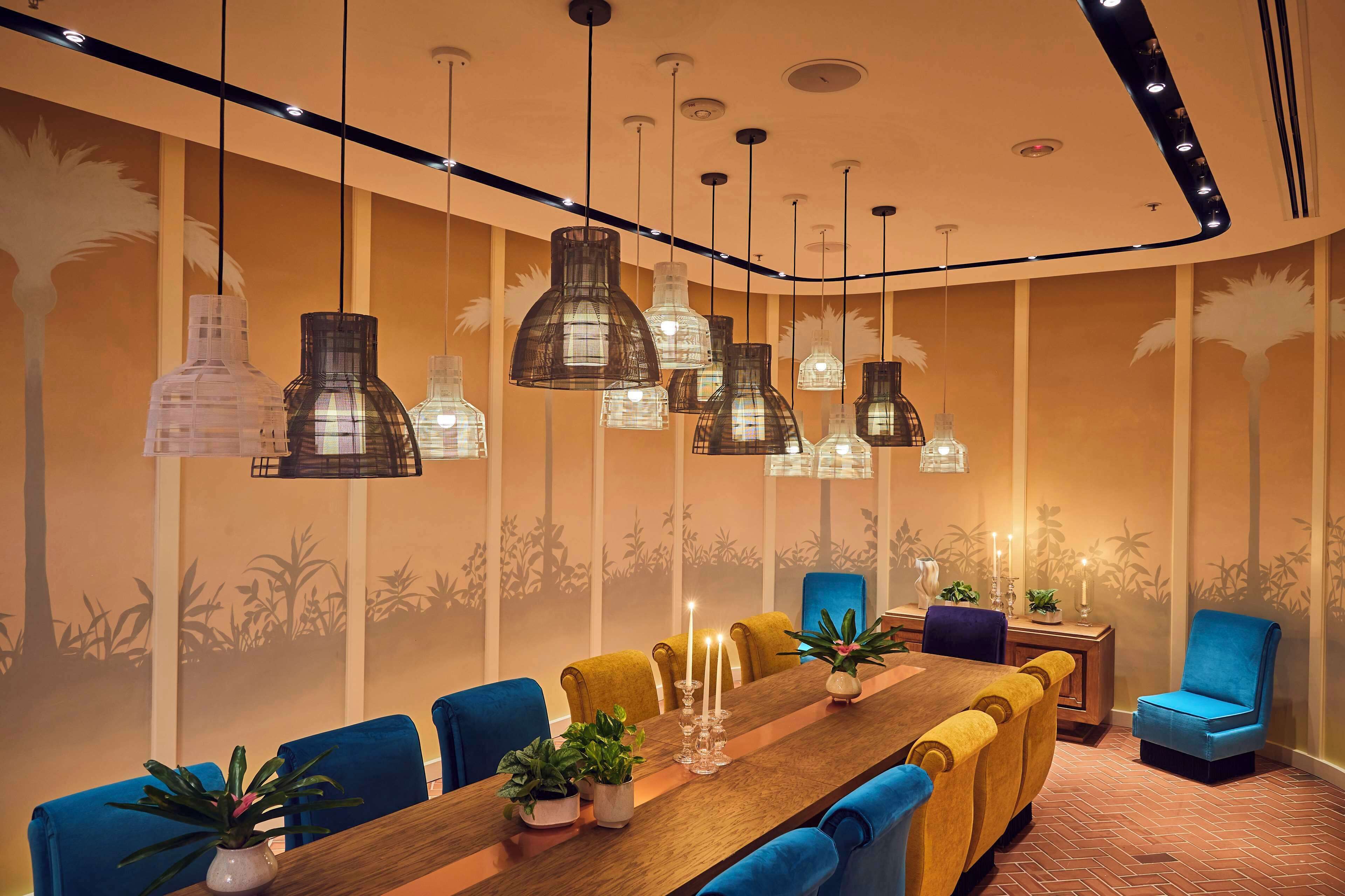 Een congreszaal met een lange houten tafel, blauwe en gele stoelen, hangende吊灯 en muurschilderingen.