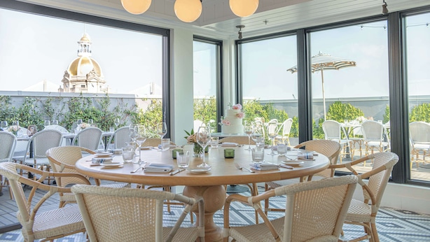 Un comedor con mesas redondas y sillas de mimbre, ventanas amplias con vista a un edificio con cúpula.