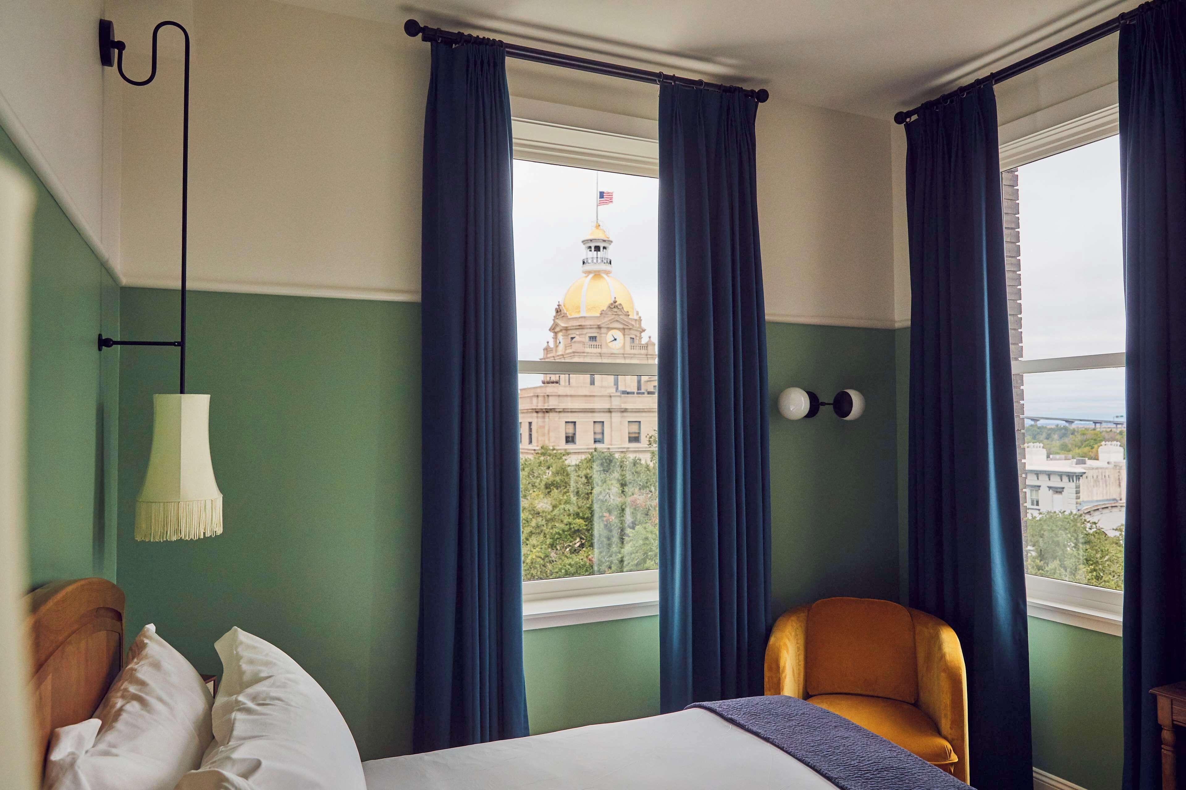 A hotel room with a bed, a chair, a lamp, and a view of a building with a dome.
