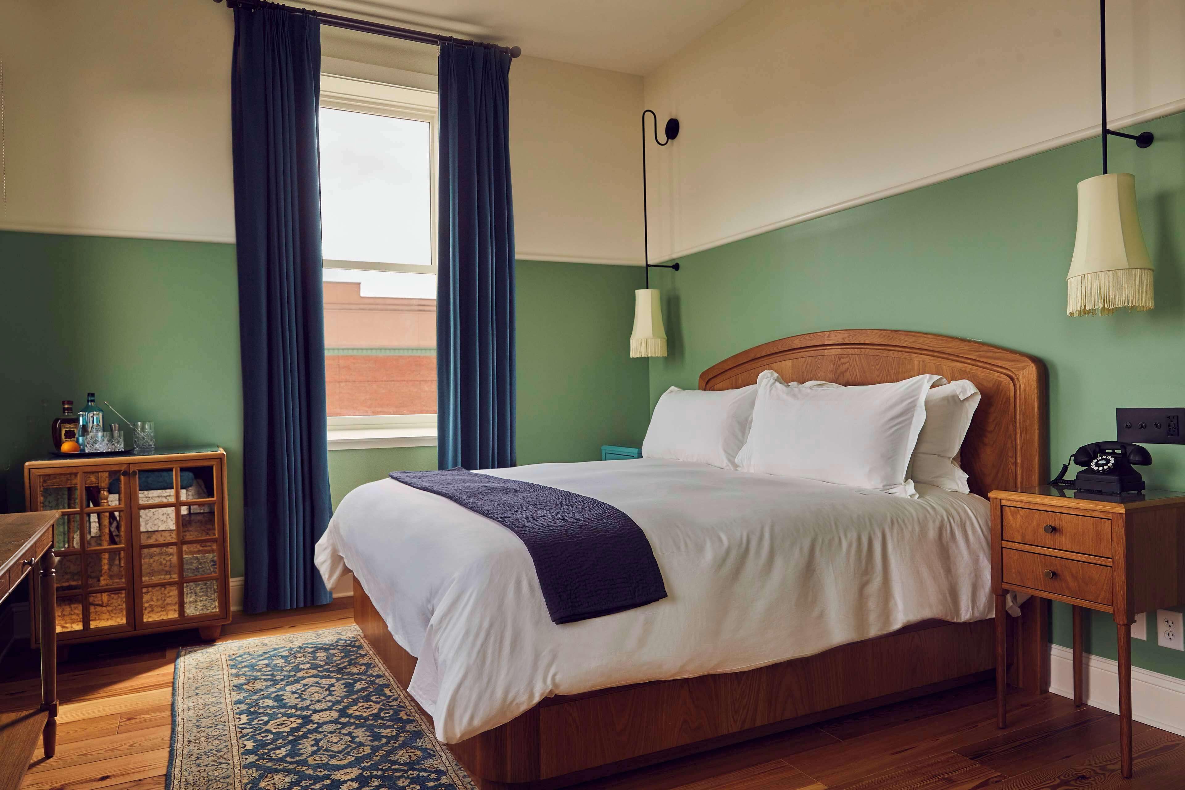 A hotel room with a bed, bedside table, lamp, and a view of the outside.