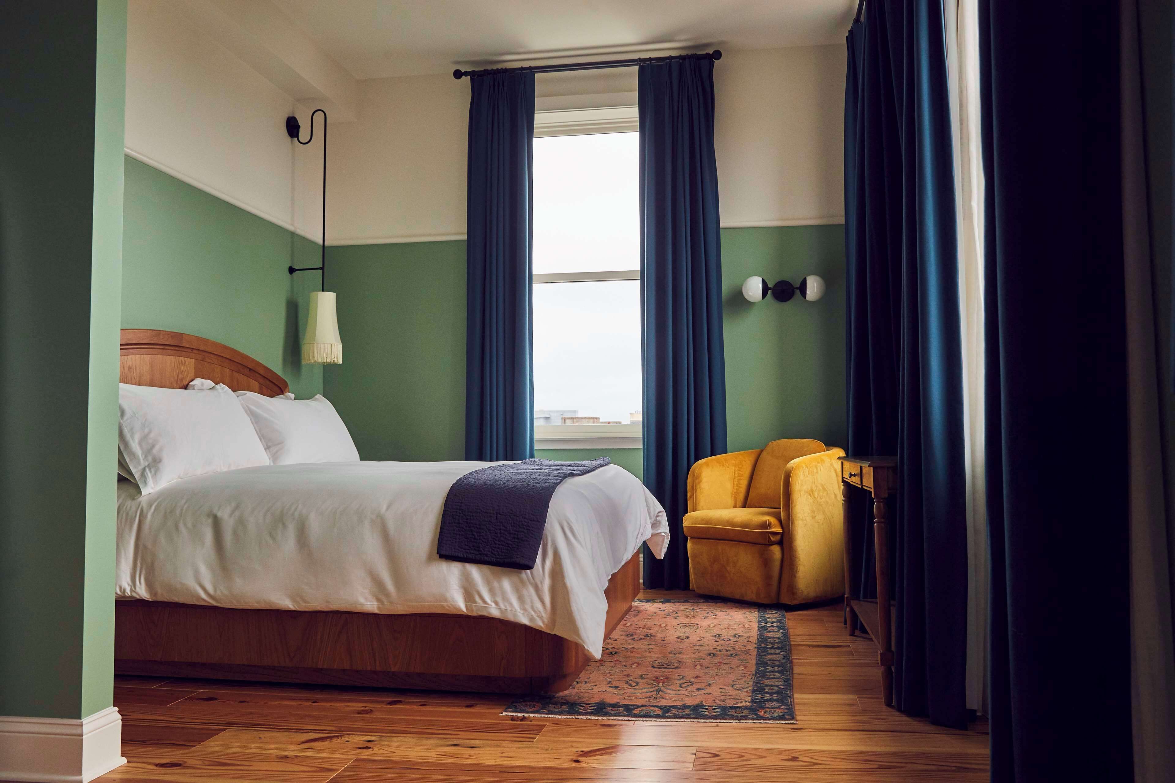 A hotel room with a bed, a chair, a window with curtains, and a lamp.