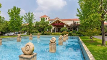 Una piscina con caracolas decorativas, un edificio con techo rojo y vegetación circundante.