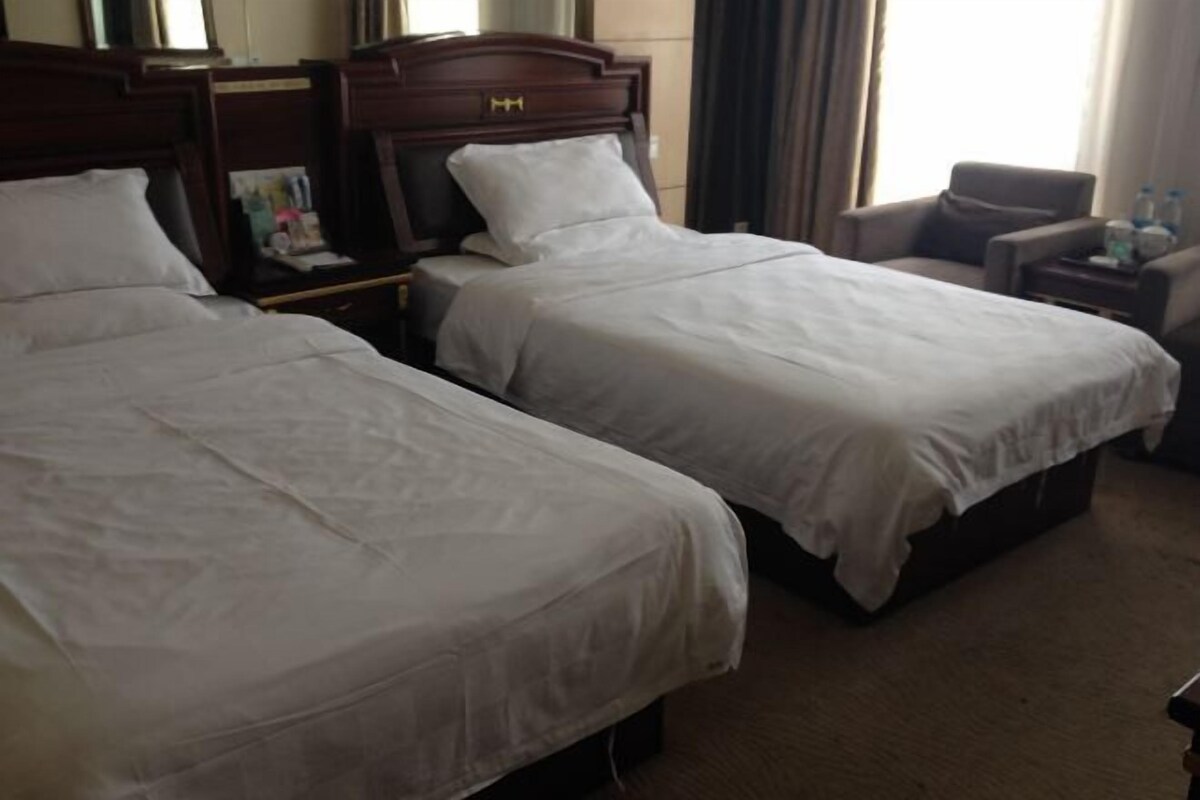 A hotel room with two single beds, a wooden headboard, a nightstand, a chair, and a small table with water bottles.