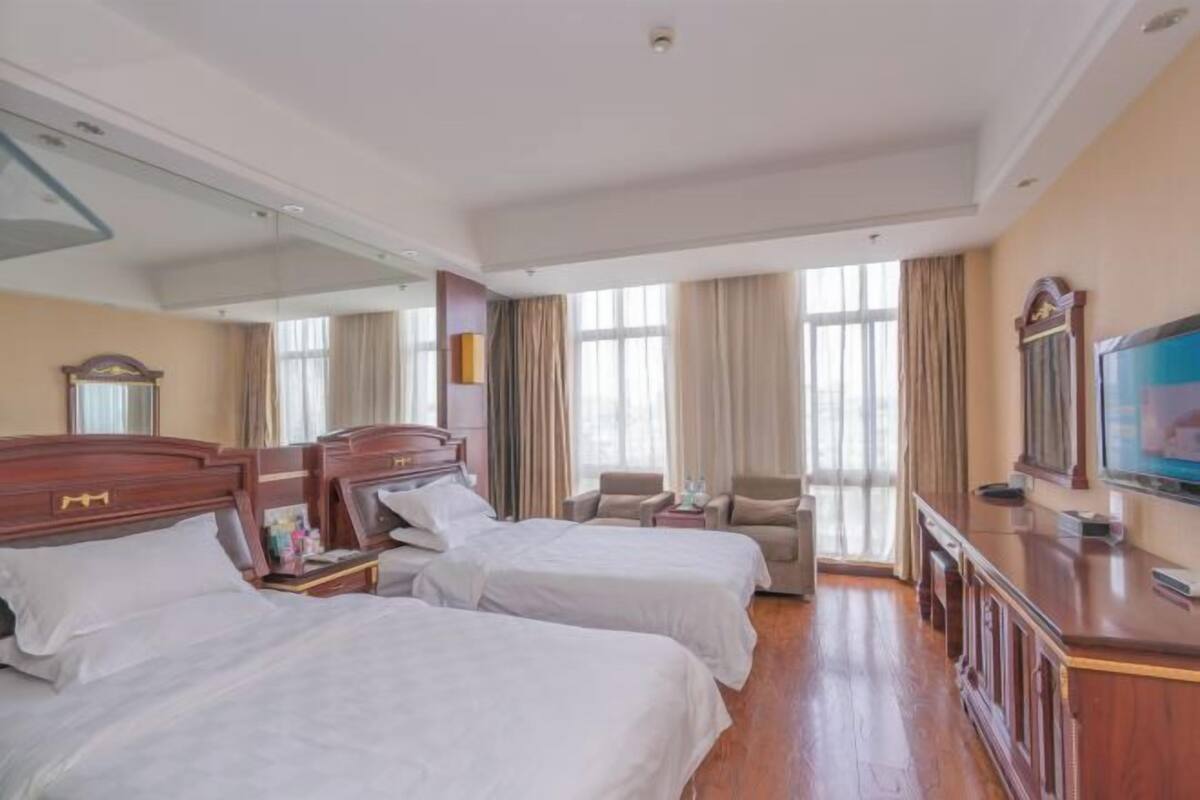 A hotel room with two beds, a television, a desk, and a large window with curtains.