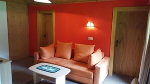 Una habitación con una pared roja, una puerta de madera, un sofá beige con cojines naranja y una mesita blanca con un objeto decorativo.