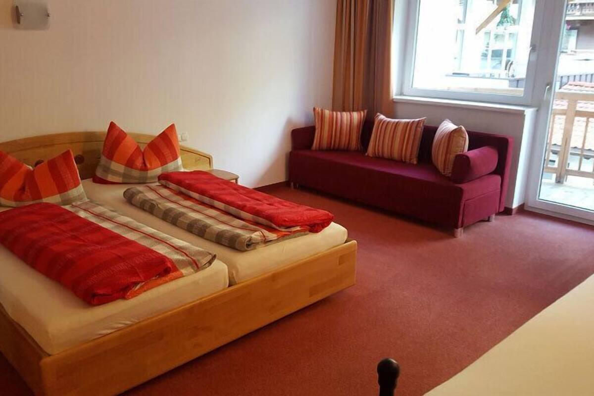 A room with a wooden bed, red and striped pillows, a red blanket, a wooden nightstand, a red sofa with striped cushions, and a window with curtains.