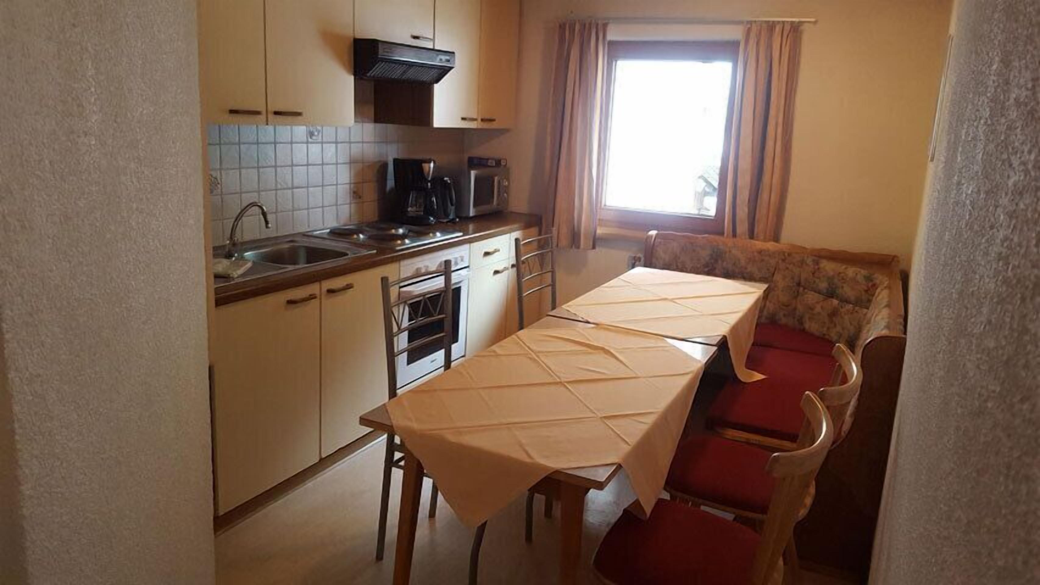 A compact kitchen with a dining area, including a table with a tablecloth, chairs, and a window with curtains.