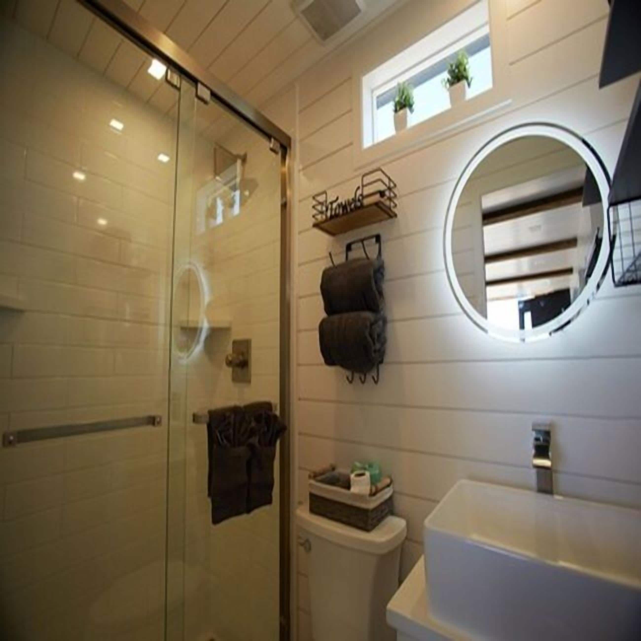 Un baño con cabina de ducha de vidrio, un lavamanos blanco, un espejo redondo y una pequeña ventana con plantas.