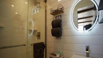 Un baño con cabina de ducha de vidrio, un lavamanos blanco, un espejo redondo y una pequeña ventana con plantas.