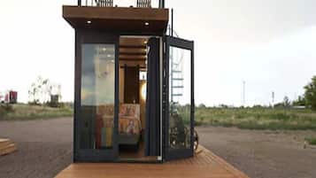 A compact, modern structure with a glass door entrance, a small deck area, and a visible interior with a bed and seating.