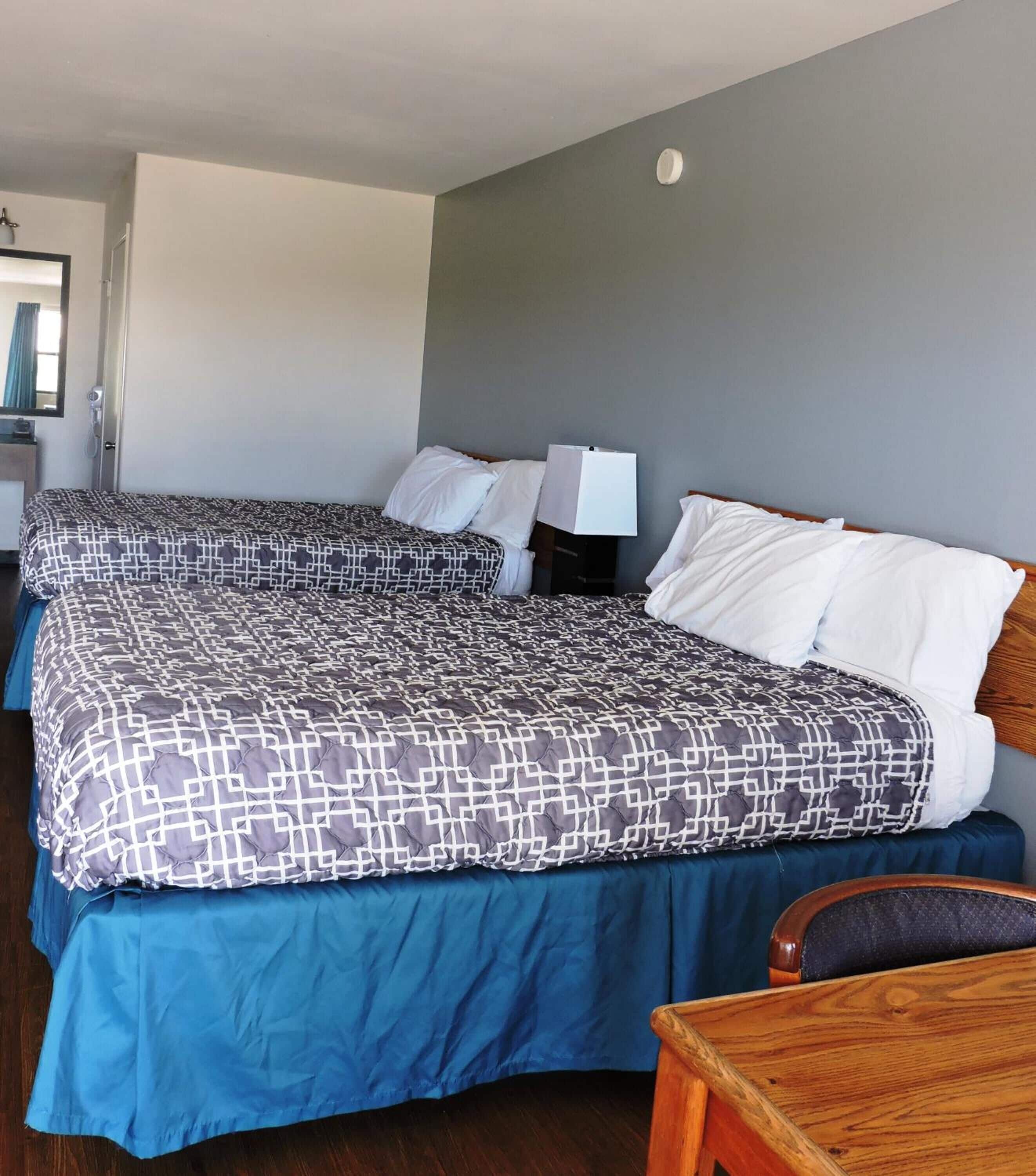Une chambre d’hôtel avec deux lits, chacun recouvert d’une couvre-lit à motifs et doté d’oreillers blancs. On y trouve une chaise en bois et une table de chevet avec une lampe.