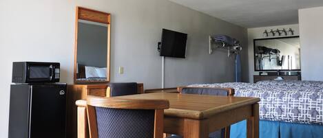 A hotel room with a wooden dining table, two chairs, a bed with a patterned bedspread, a flat-screen TV mounted on the wall, a microwave on a small appliance, and a large mirror above the bed.