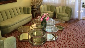 Un salon avec un canapé vert, deux fauteuils, une table basse en verre ornée d’un vase de fleurs, un lustre et un tableau accroché au mur.