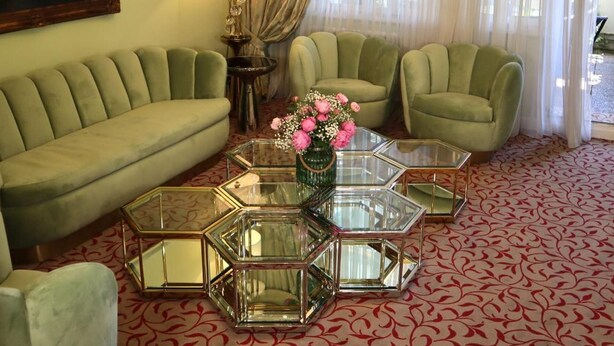 Un salón con un sofá verde, dos sillones, una mesa de centro de vidrio con un florero y una lámpara de araña, además de un cuadro en la pared.