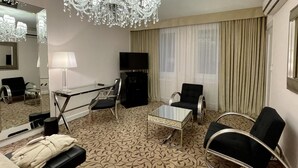 Habitación de hotel con candelabro, televisor de pantalla plana, mesa de centro de vidrio, dos sillones, un sofá y un espejo grande.