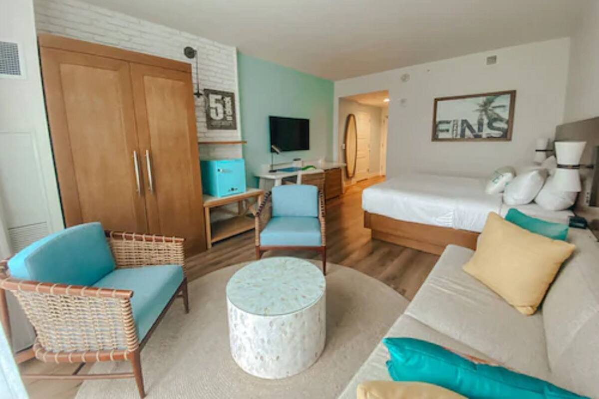 A hotel room with a bed, a sofa, a chair, a small table, a wardrobe, a television, and a painting on the wall.