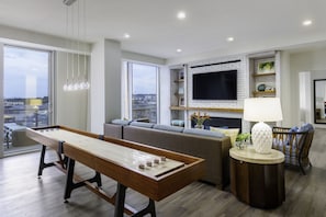 A modern living room with a foosball table, a grey sofa, a TV mounted on a brick wall, and a large window offering a city view.