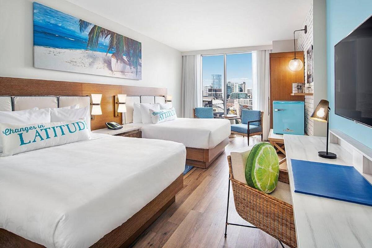 A hotel room with two beds, a city view, a painting of a beach, and a large TV.