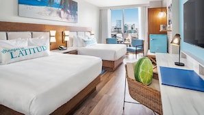 Une chambre d’hôtel avec deux lits, une vue sur la ville, un tableau représentant une plage et un grand téléviseur.