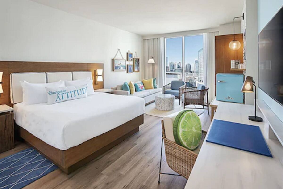 A hotel room with a large bed, a sofa, a dining table, a TV, and a city view.