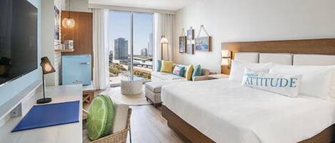 A hotel room with a large bed, a desk with a computer, a chair, a small table with a watermelon, and a view of the city through the window.