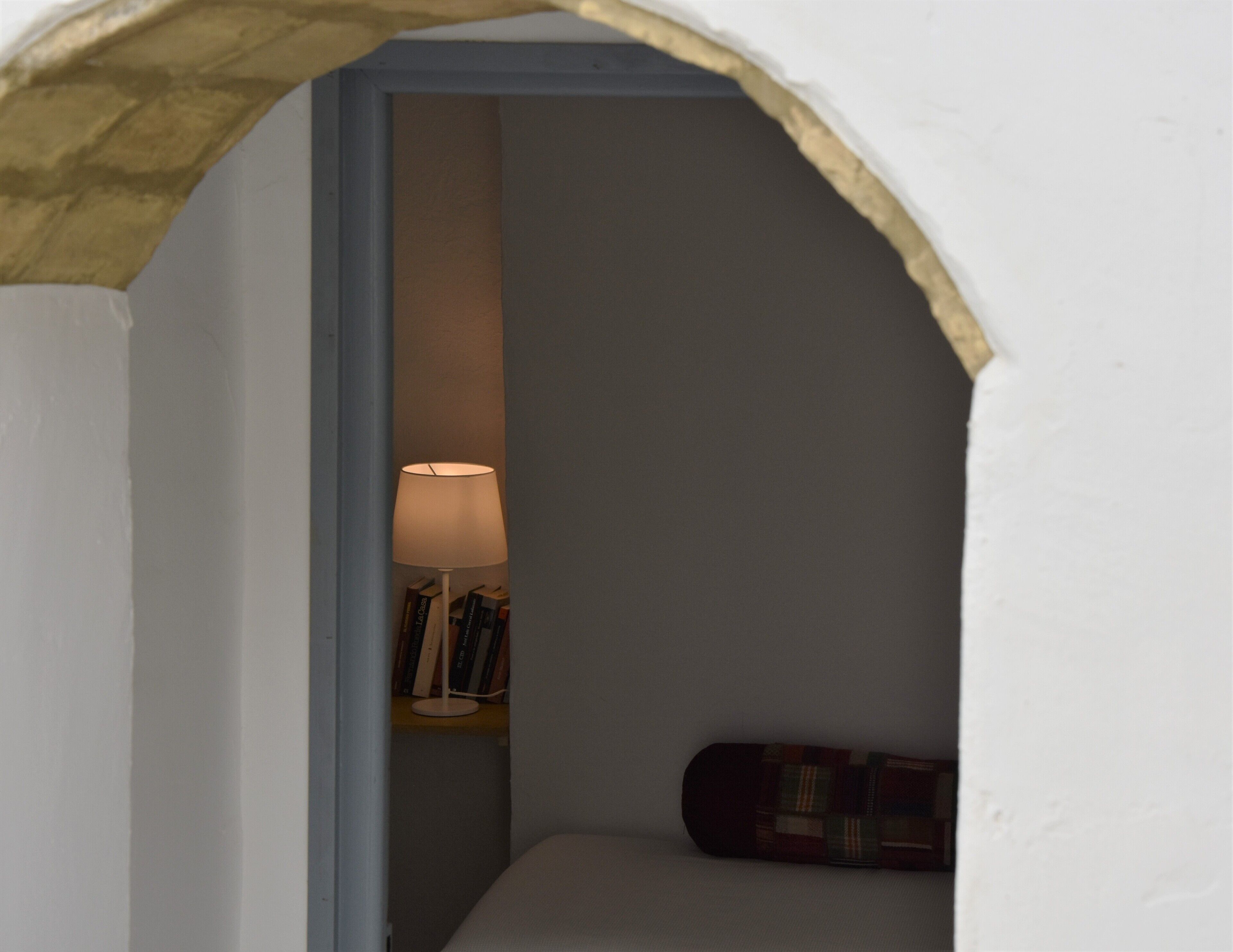 Un estrecho arco de entrada que conduce a una pequeña habitación con una cama, una lámpara y una estantería llena de libros.
