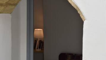 Un estrecho arco de entrada que conduce a una pequeña habitación con una cama, una lámpara y una estantería llena de libros.