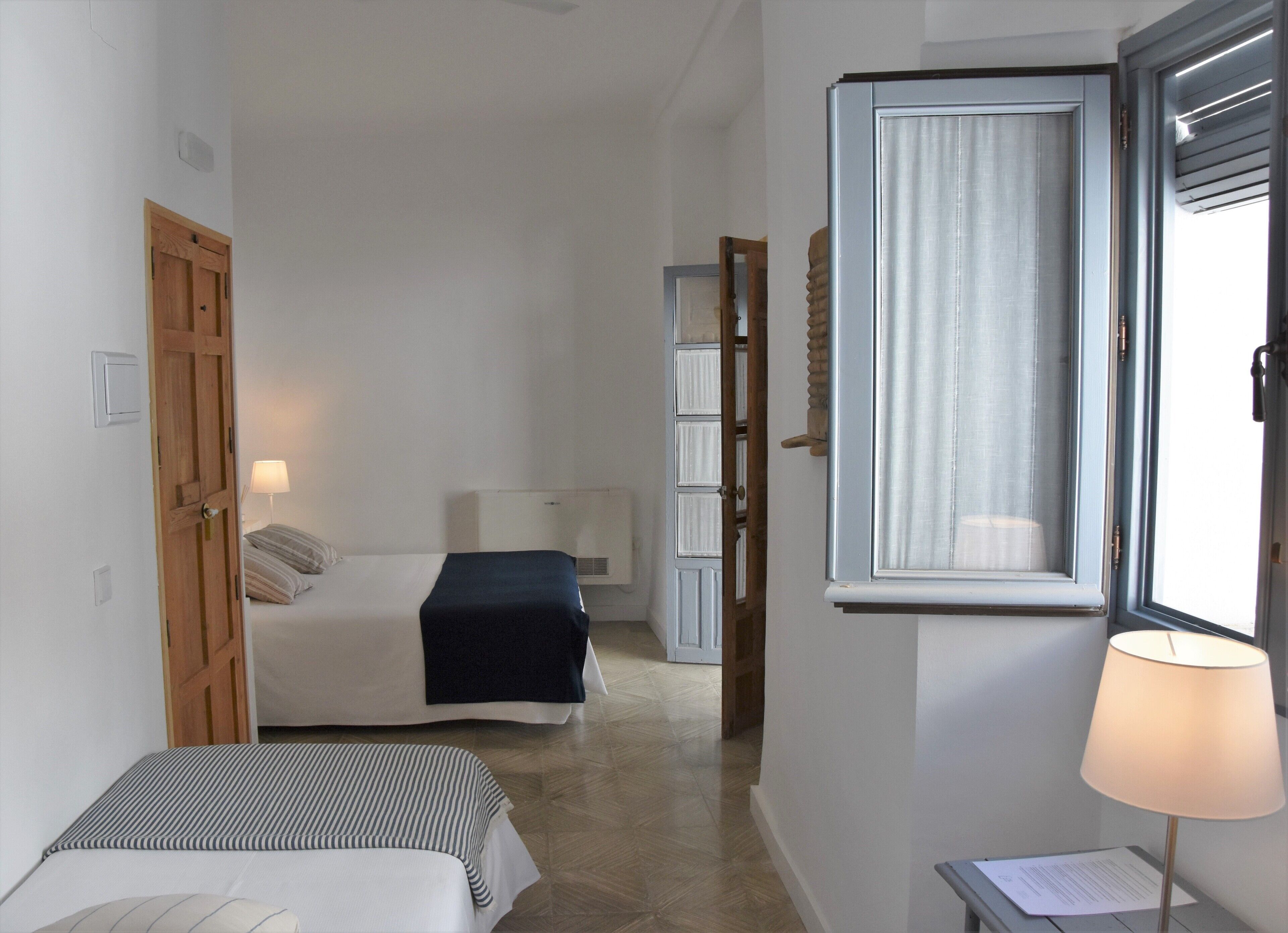 Kapea hotellihuone, jossa on kaksi sänkyä, ikkuna, jossa on verhot, yöpöytä lampun kanssa ja puinen ovi.