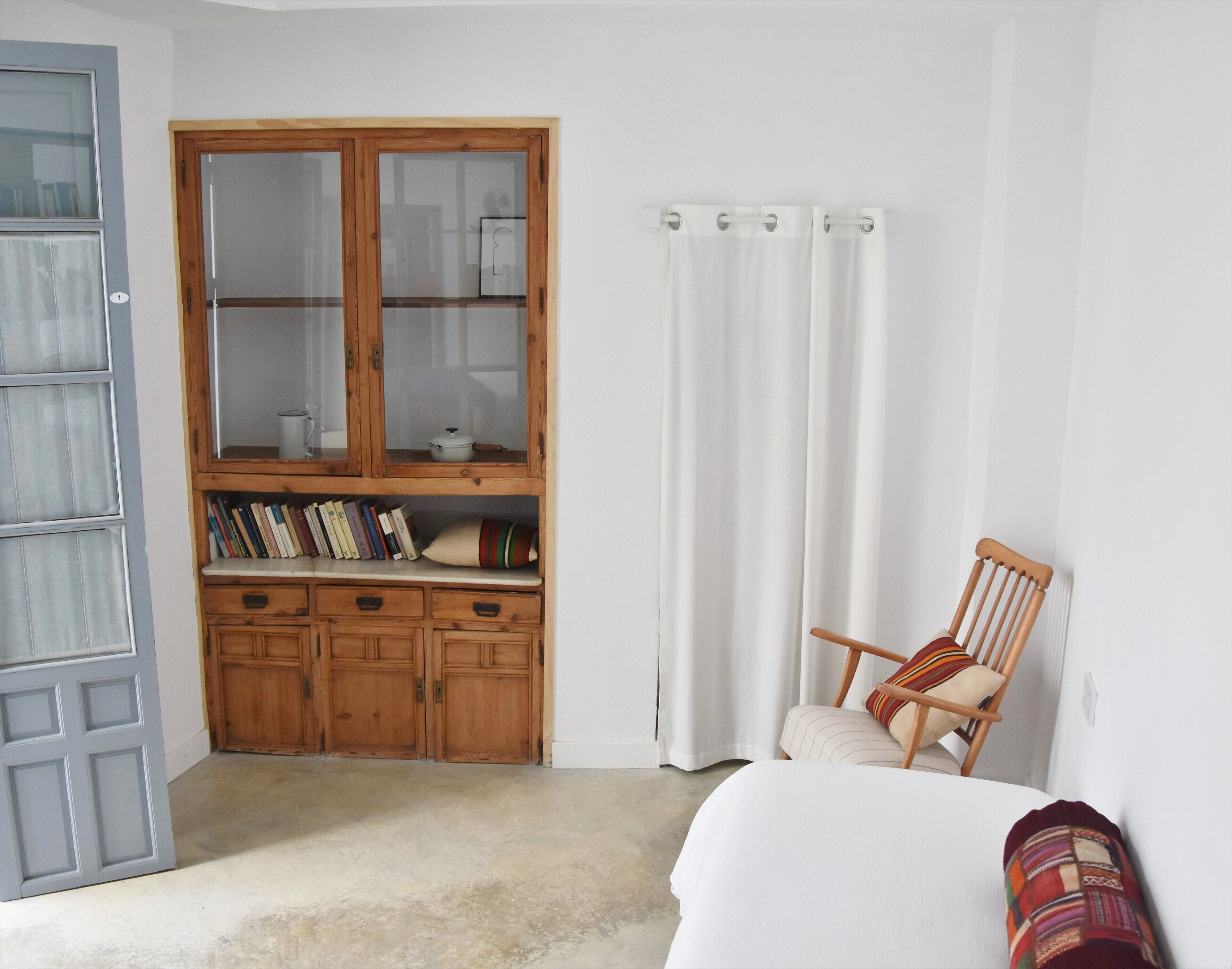 Un dormitorio con un armario de madera, una silla, una cama y un ventanal con cortinas.