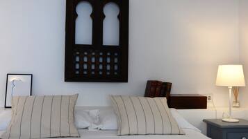 Een opgemaakt bed met twee kussens, een wanddecoratie, een nachtkastje met een lamp en een bijzettafeltje met boeken.