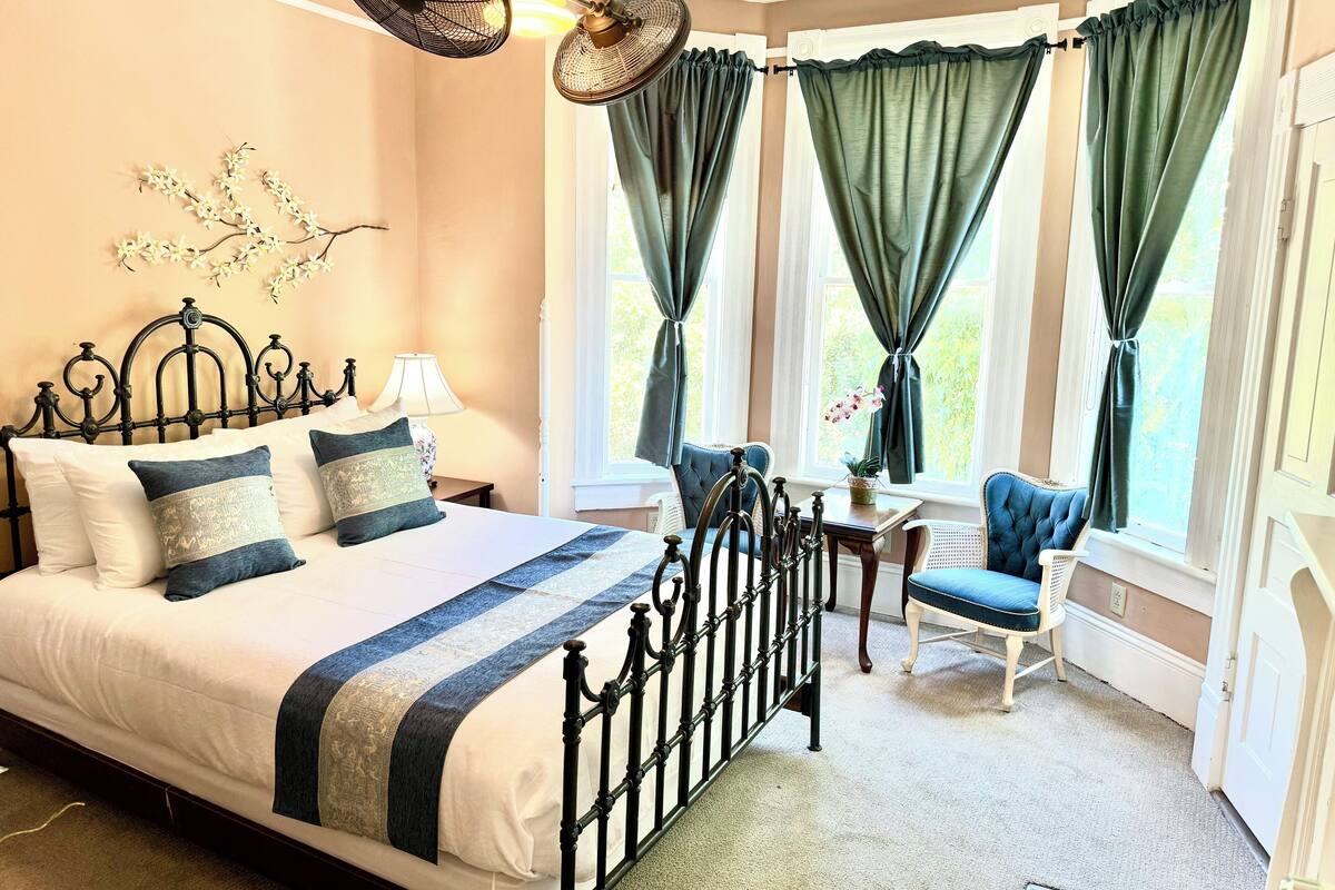 Quarto com cama grande, duas cadeiras, uma mesa e janela com cortinas.