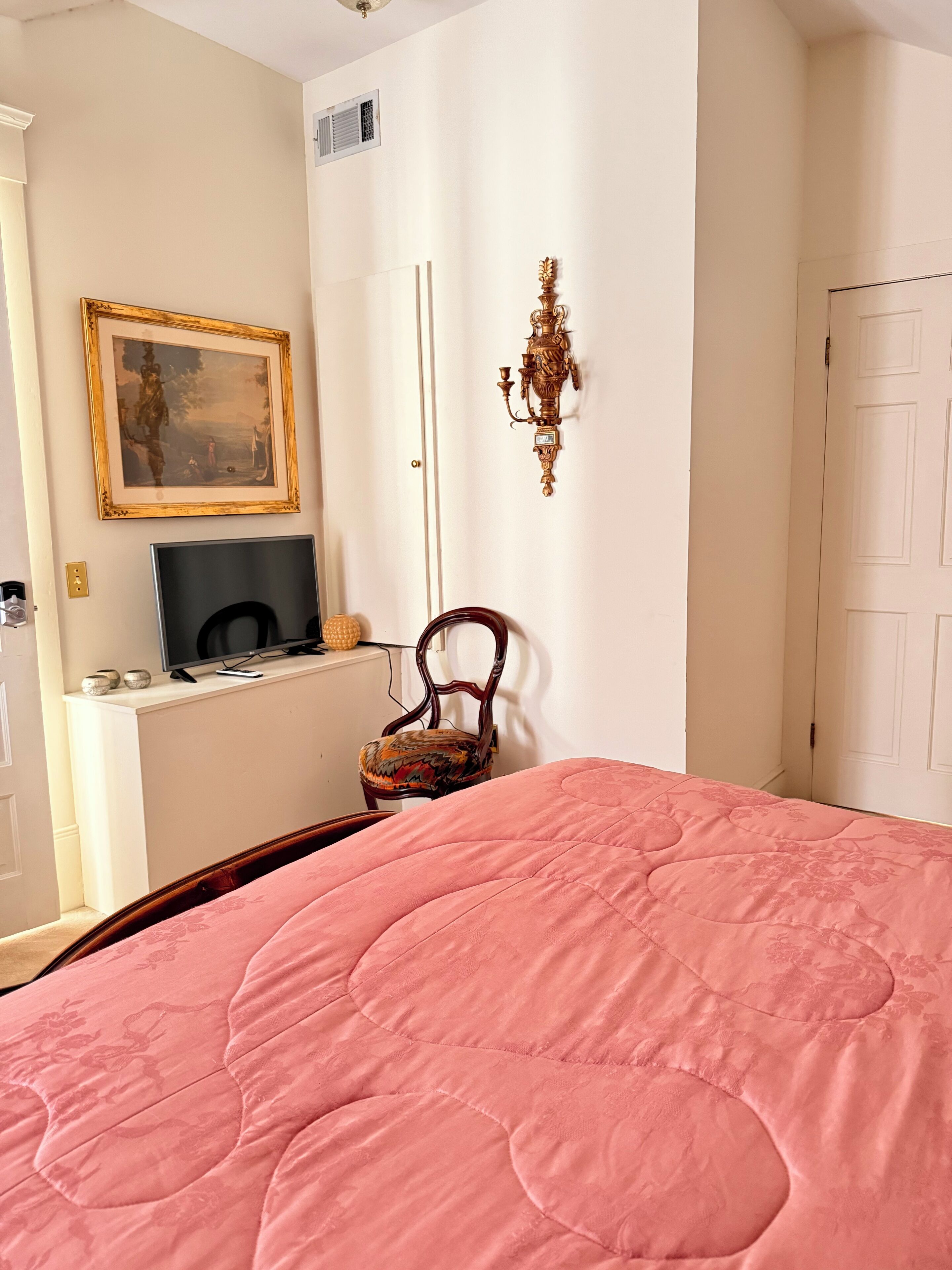 A bedroom with a pink quilted bedspread, a wooden chair, a television on a white stand, a framed painting, and a decorative wall sconce.