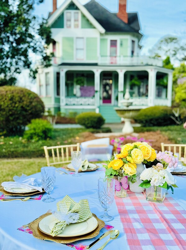 A dining table set outdoors with a view of a green house in the background.