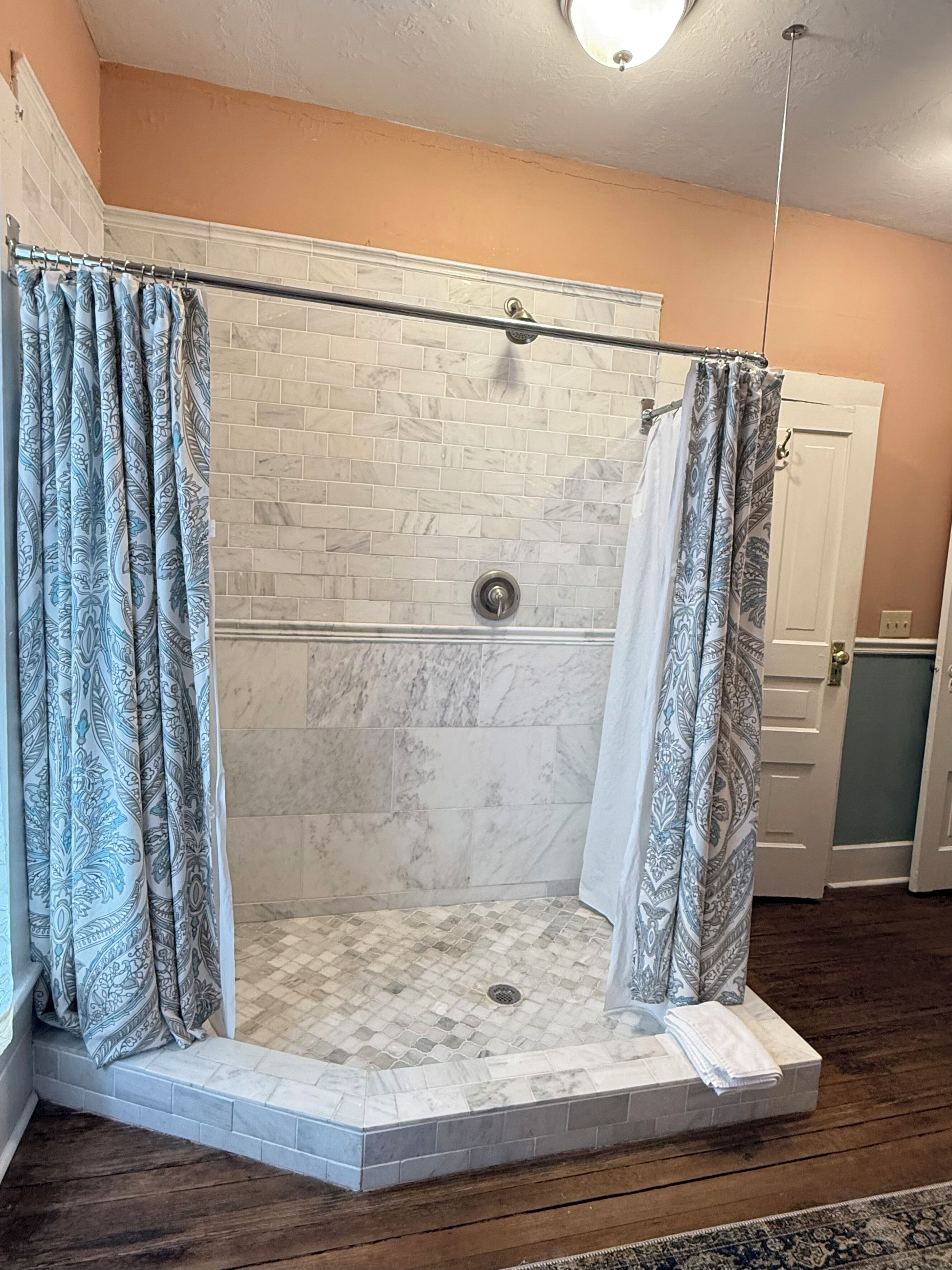 A walk-in shower with patterned curtains, a tiled floor, and a white door in the background.