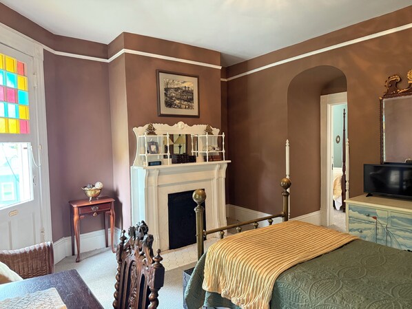 A bedroom with a fireplace, a bed, a television, and a window with colorful stained glass.